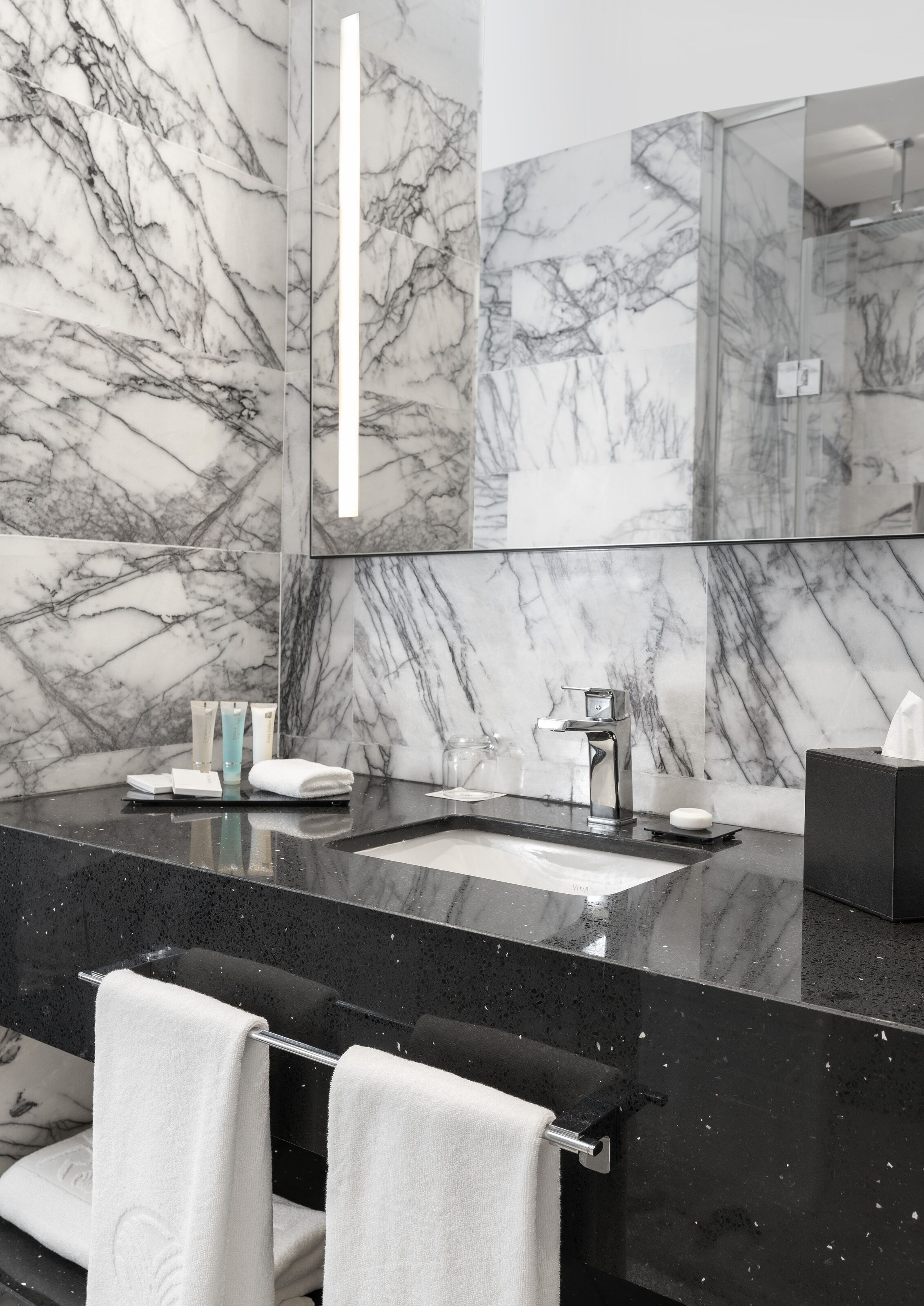A modern bathroom with a marble backsplash, a black countertop, a sink, a mirror, and two towels on a rack.
