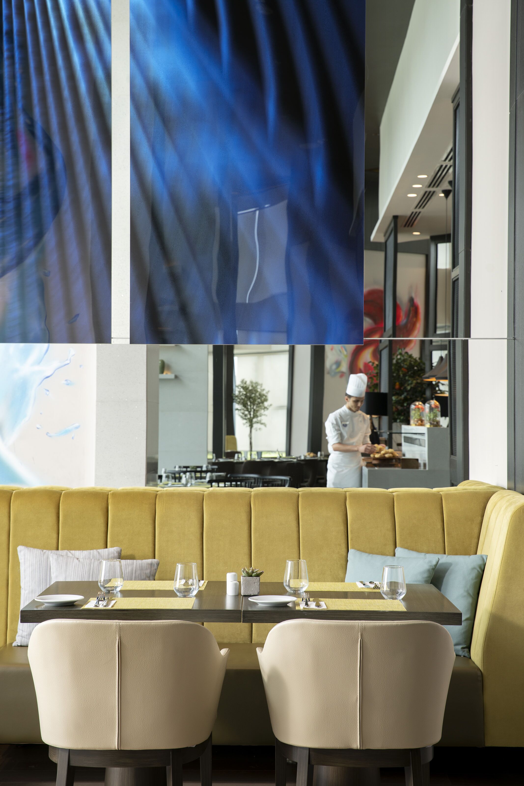 A modern dining area with a yellow booth and two chairs, a table set with glasses and cutlery, and a chef in the background.
