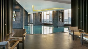 A modern indoor swimming pool with a view of the cityscape, featuring stone walls, wooden furniture, and a clear pool. - Westside Residences By Rotana (Istanbul)