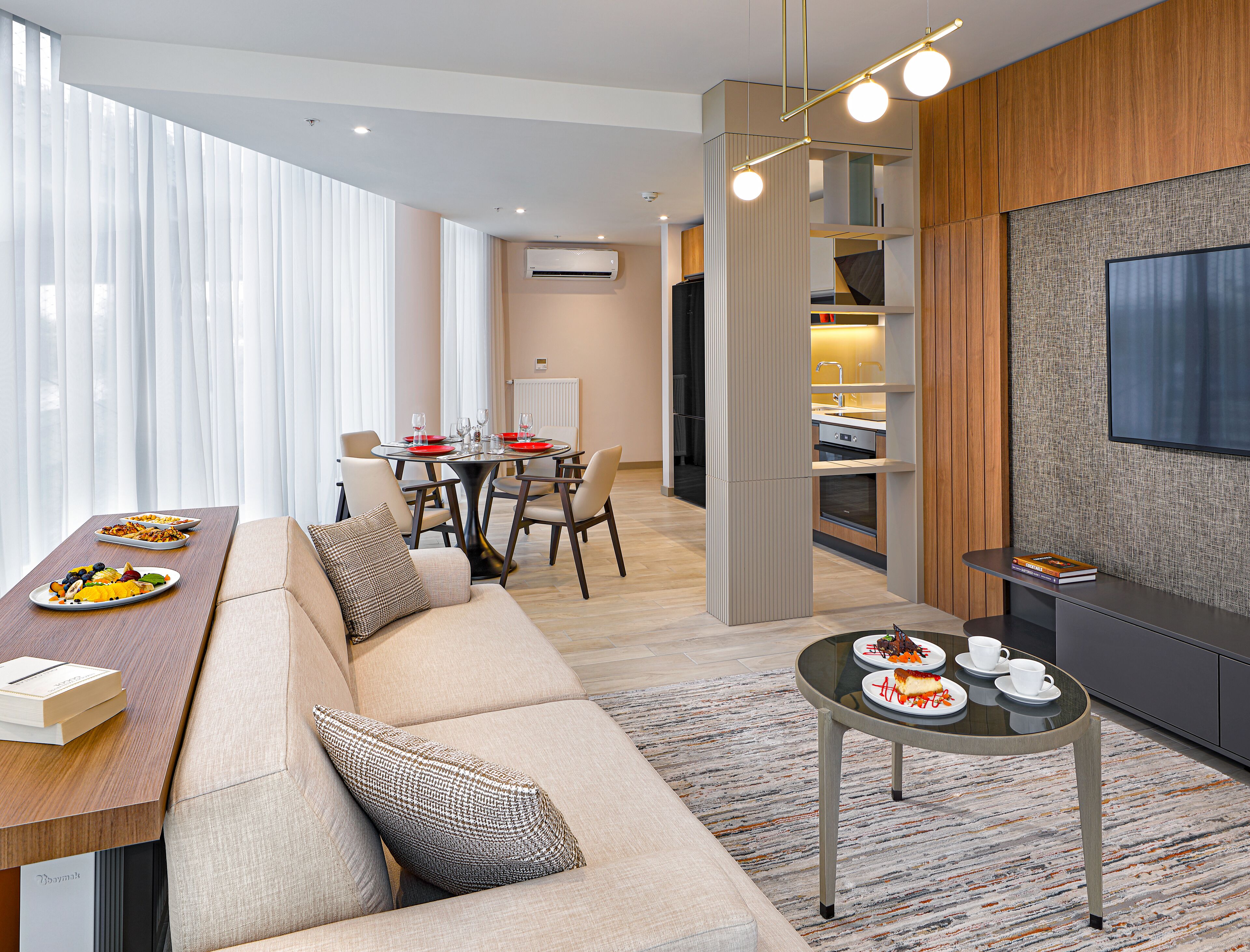 A modern hotel room with a dining area, a sofa, a television, and a kitchen area.