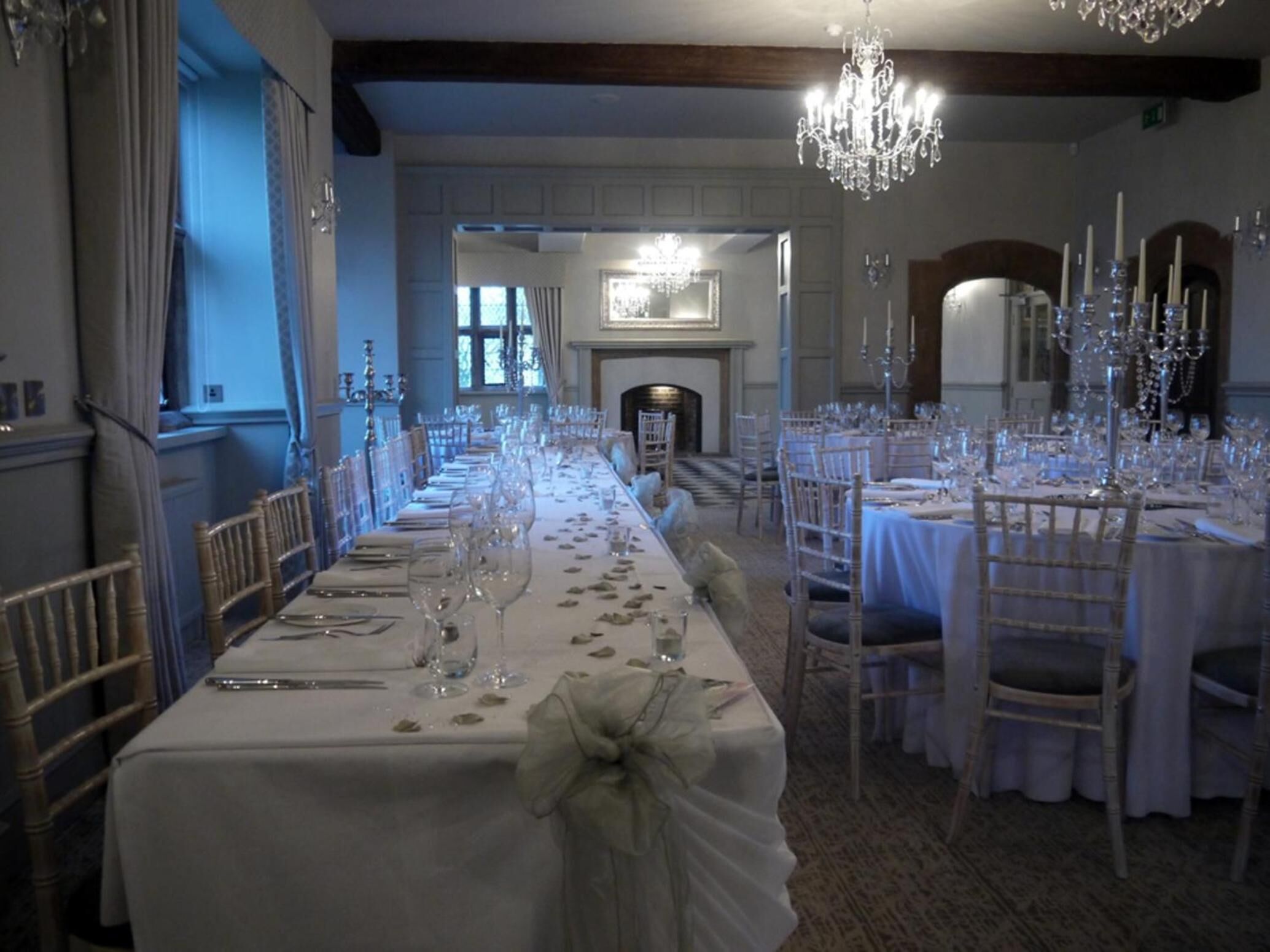 A formal dining room with long tables set for a meal, chandeliers, and a fireplace in the background.