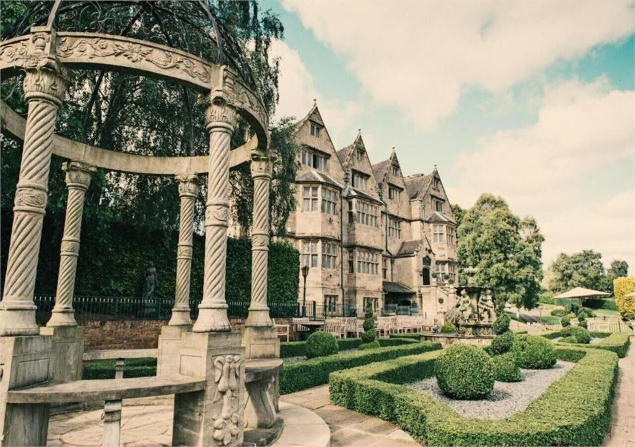 A historic building with a symmetrical facade, a central entrance with a decorative archway, and a well-manicured garden with hedges and a fountain.