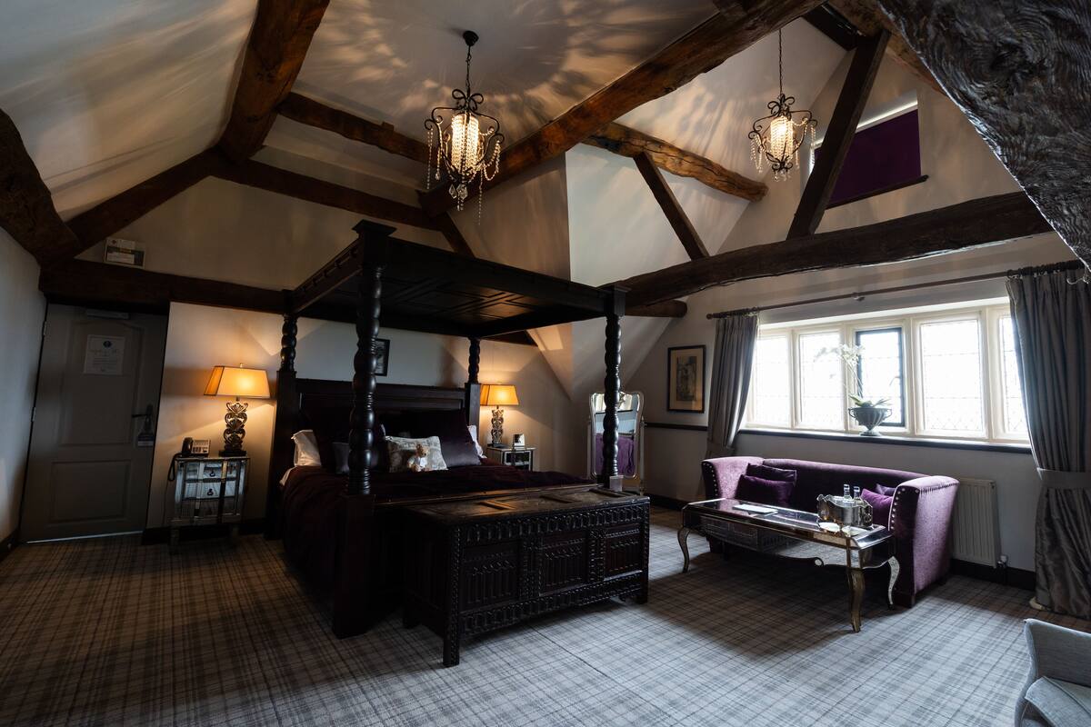 A four-poster bed with a wooden headboard, a sitting area with a table and chairs, and a large window with curtains.