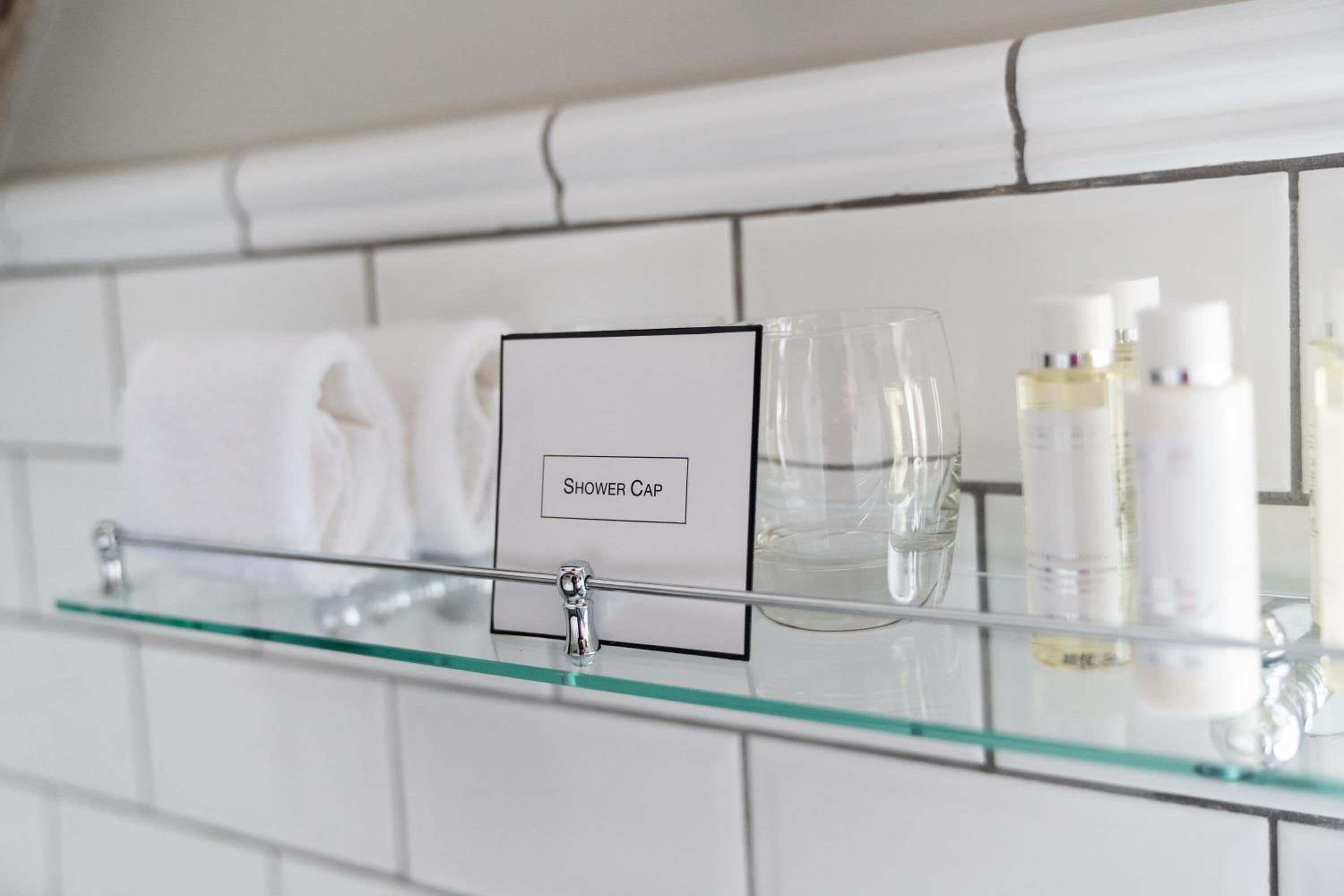 Une salle de bain avec une étagère en verre sur laquelle sont posés un bonnet de douche, un verre et des articles de toilette.