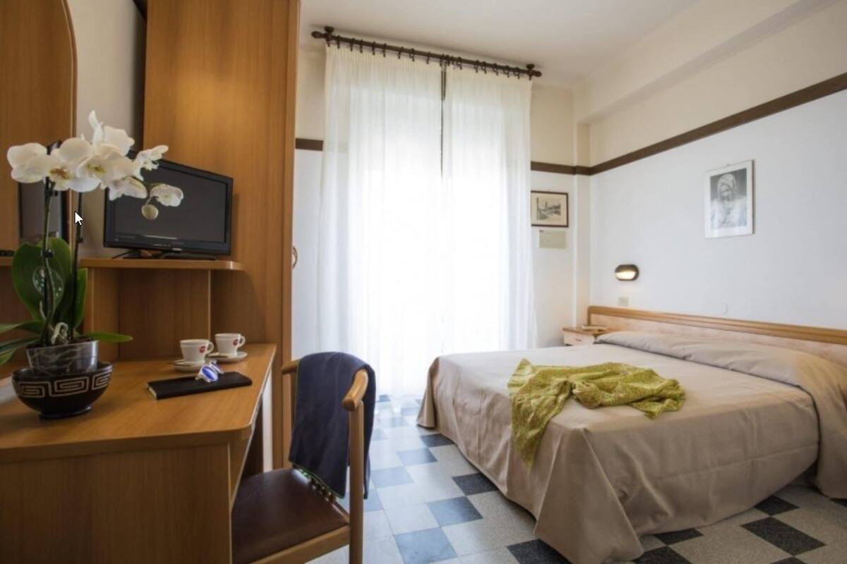 A hotel room with a bed, a desk with a computer and a vase of flowers, a chair, and a window with curtains.