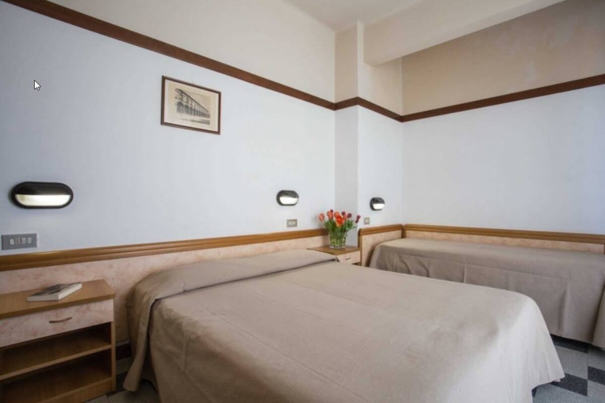 A hotel room with two beds, a nightstand, a lamp, a framed picture, and a vase with flowers.