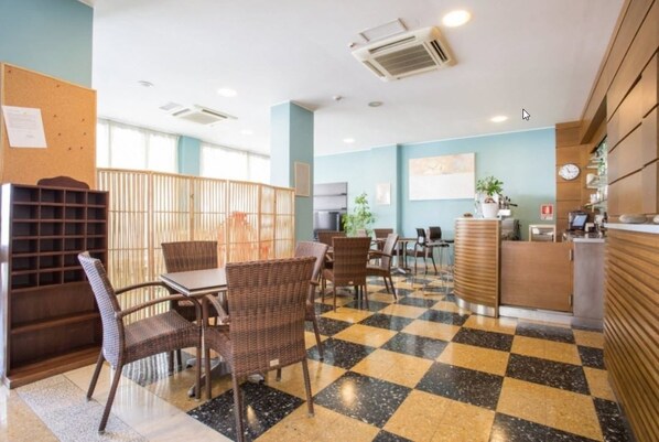 A hotel lobby with checkered flooring, wicker furniture, a reception desk, and a bulletin board.