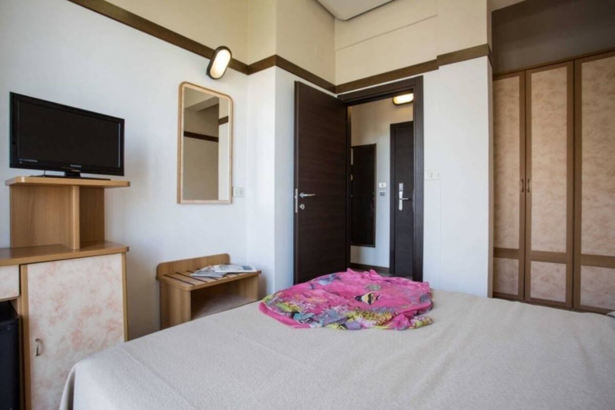 A hotel room with a bed, a television, a mirror, a wardrobe, and a small table with a chair.