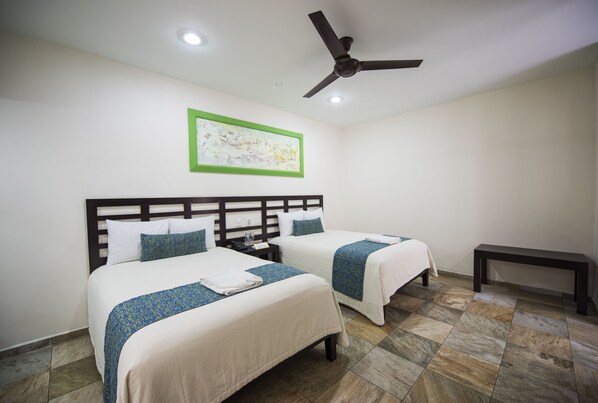 Una habitación de hotel con dos camas, un ventilador de techo, una mesita de noche, un banco y un cuadro en la pared.