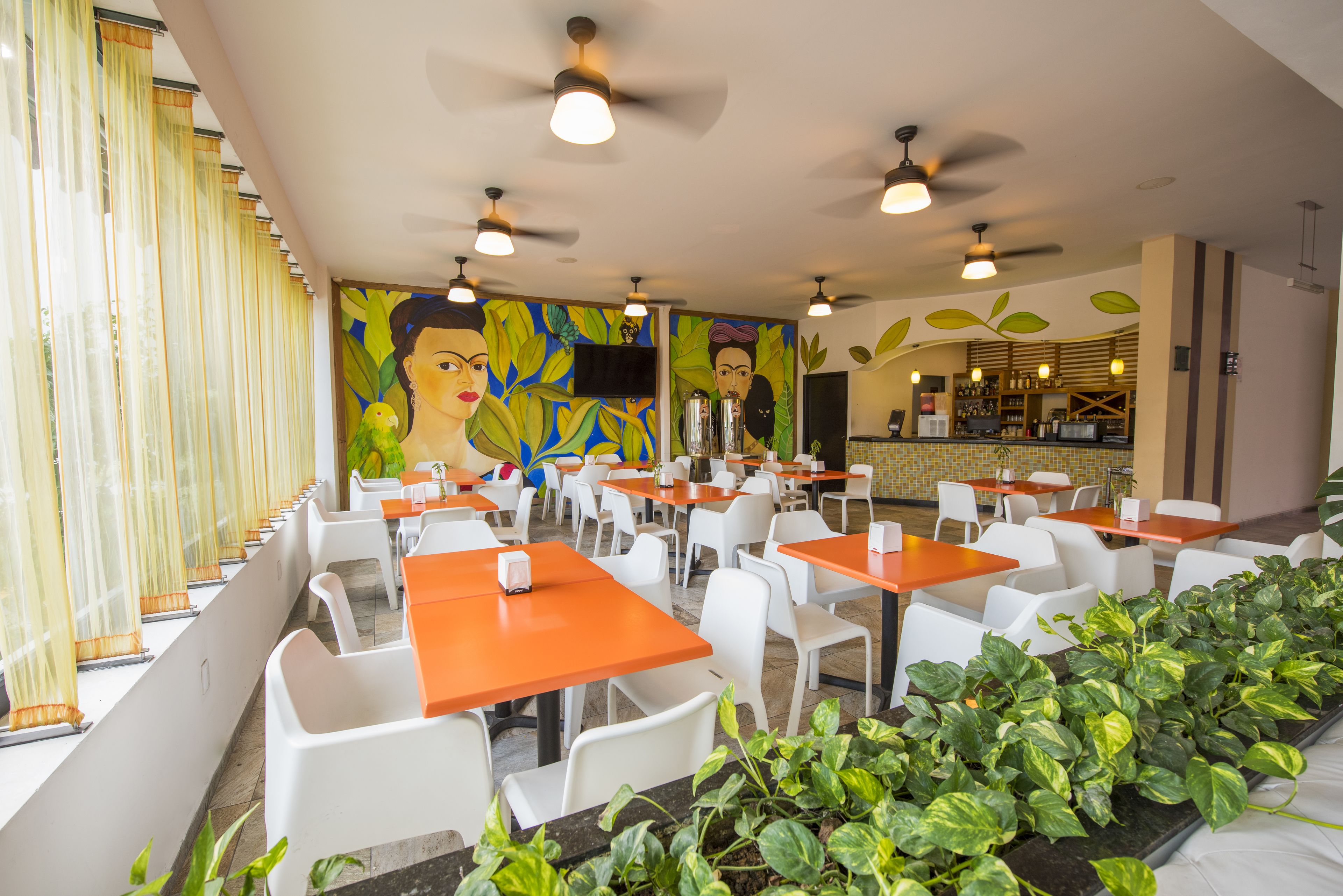 Un restaurante con sillas blancas y mesas naranjas, un mural en la pared y ventiladores de techo.