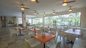 A spacious indoor dining area with multiple tables and chairs, ceiling fans, and large windows offering a view of palm trees and parked cars. - Hotel Andiroba Palace (Tuxtla Gutierrez)