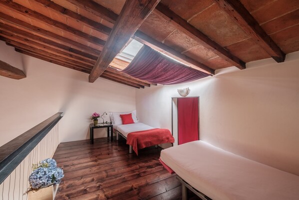 A small, cozy room with wooden beams, a sloped ceiling, a bed with a red blanket, a bedside table with a lamp, and a window with a curtain. - Porcellana Apartment (Florence)