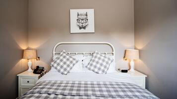 A neatly made bed with a checkered bedspread, two bedside tables with lamps, a wall-mounted telephone, and a framed picture of a cat wearing glasses.