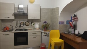 Een kleine keuken met een gasfornuis, spoelbak en een kleine koelkast. Er staat een gele stoel en een rode emmer op de vloer.