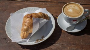 Een croissant op een met papier bekleed bord en een kop koffie met een lepel op een houten tafel.