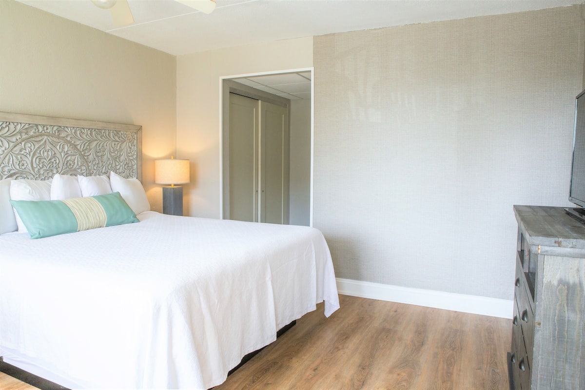 Une chambre d’hôtel avec un grand lit, une table de chevet avec une lampe, une commode en bois et une porte donnant sur une autre pièce.