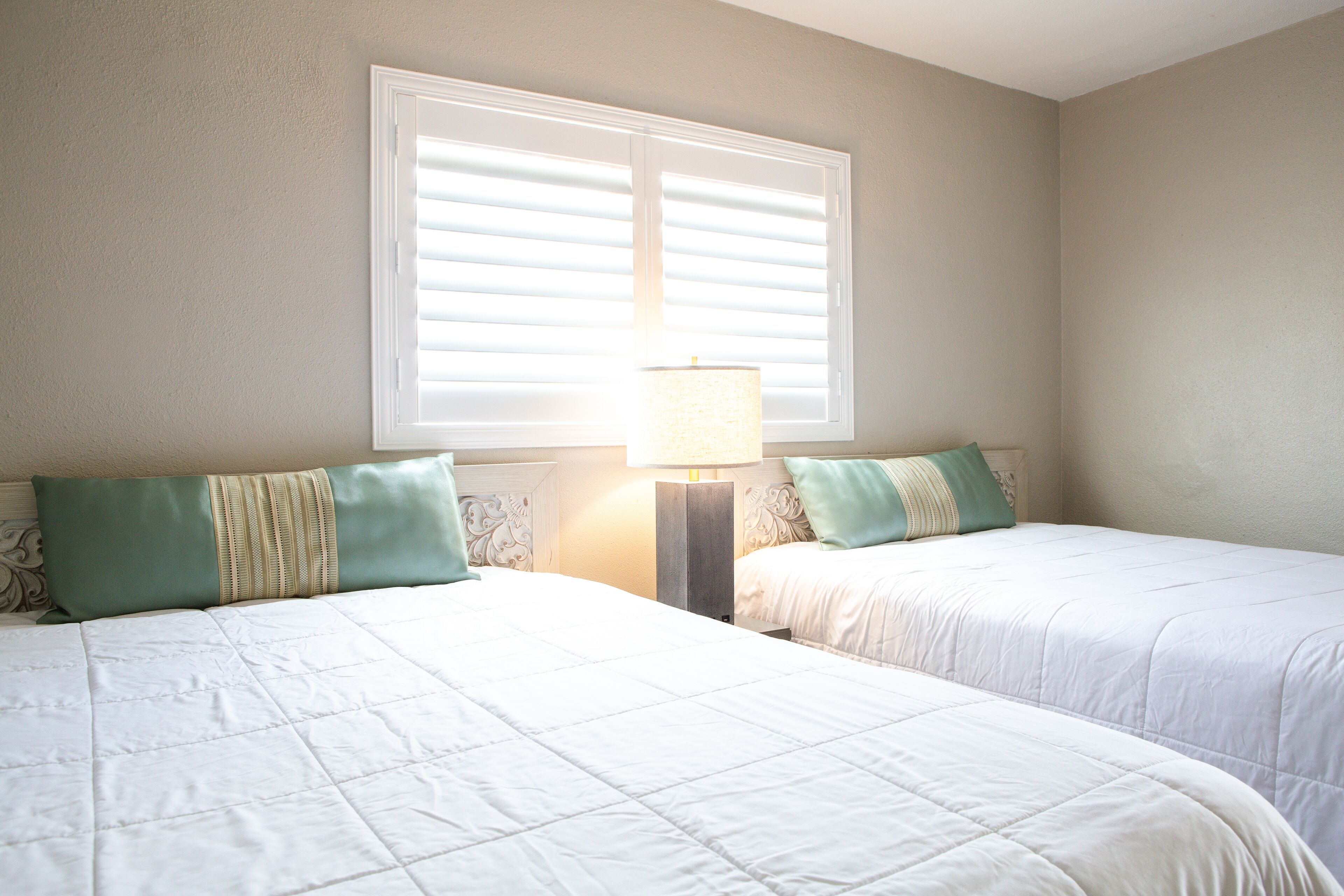 A bedroom with two beds, a window with blinds, and a bedside lamp.