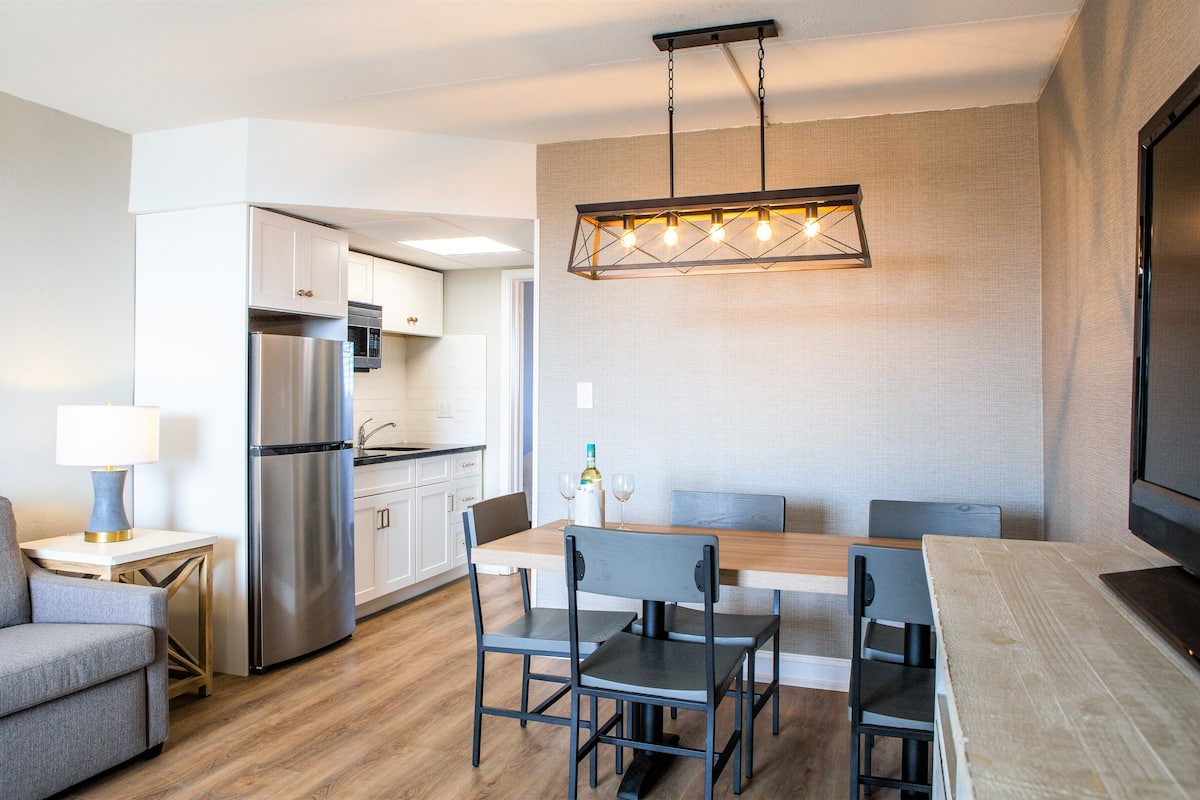 Une cuisine moderne avec un réfrigérateur en acier inoxydable, un coin repas avec une table et des chaises en bois, et un canapé gris.