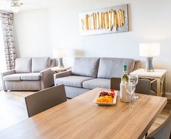 Una sala de estar moderna con dos sofás grises, una mesa de comedor de madera con una bandeja de frutas, copas de vino y una botella.