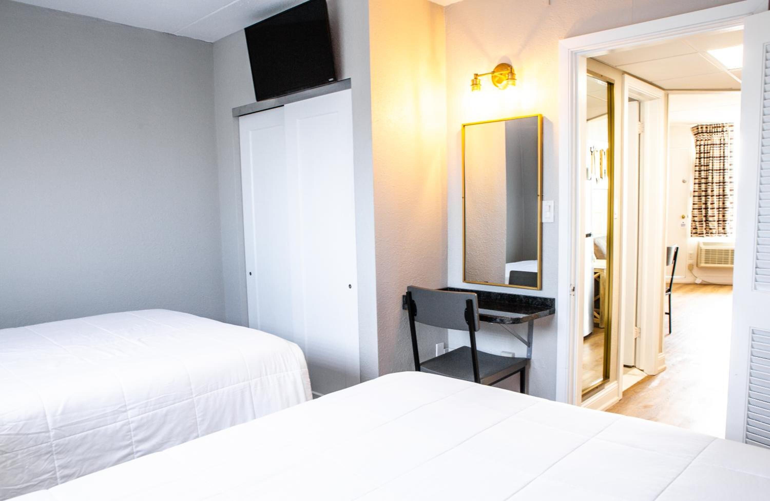 A bedroom with two beds, a desk with a chair, a mirror, and a door leading to a bathroom.