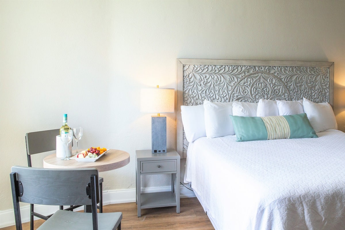 Une chambre d’hôtel avec un lit bien fait, une petite table sur laquelle se trouvent une bouteille et des verres, une chaise et une table de chevet avec une lampe.