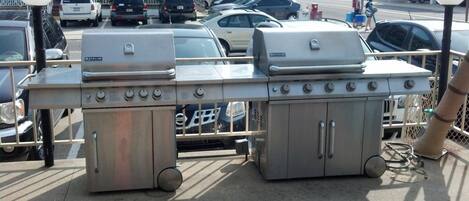 En bærbar grill i rustfrit stål med flere brændere og en indbygget ryger, placeret på en betonoverflade i nærheden af en parkeringsplads med biler.
