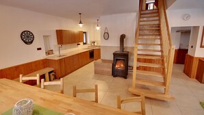 A modern kitchen with wooden cabinets, a dining area with a wooden table and chairs, a dartboard, a clock, and a wood-burning stove. - Chalupa Jiřík (Volary)