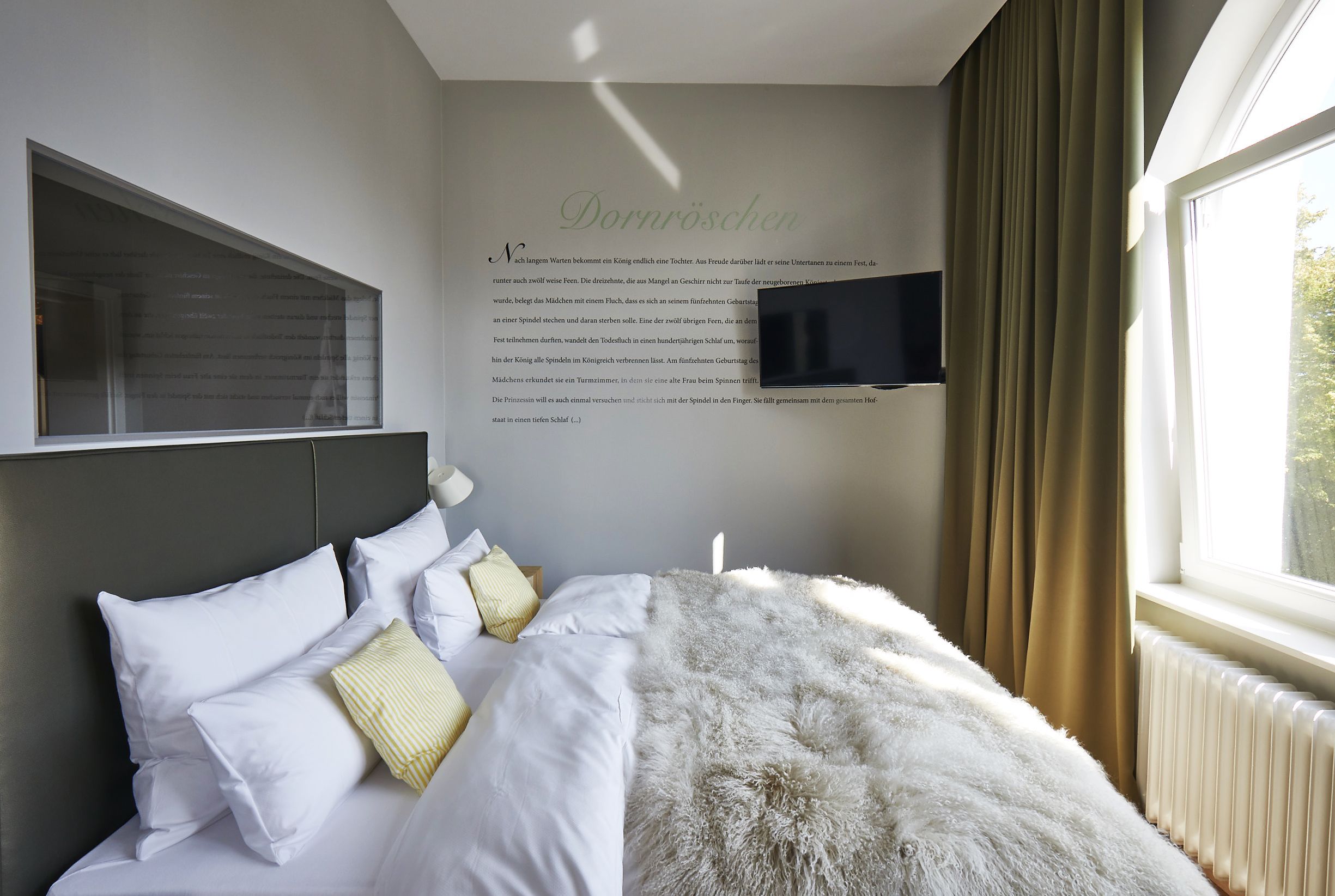A neatly made bed with white linens and a fluffy white throw blanket, a mounted flat-screen TV on the wall, and a window with curtains.