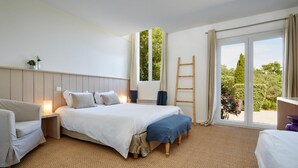 A bedroom with a large bed, a bedside table, a chair, and a view of greenery through a sliding door. - La Maison de la Prade (Messanges)