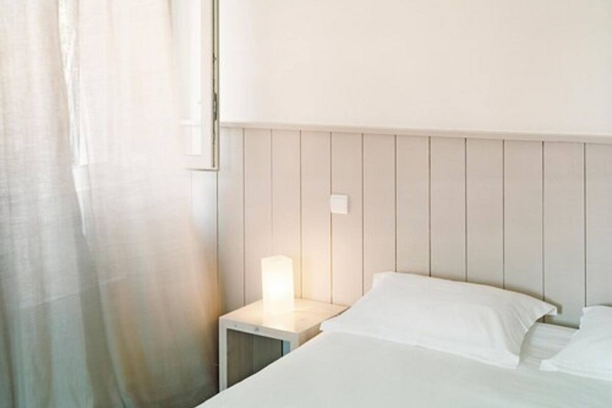 A hotel room with a bed, a nightstand with a lamp, and a window with sheer curtains.