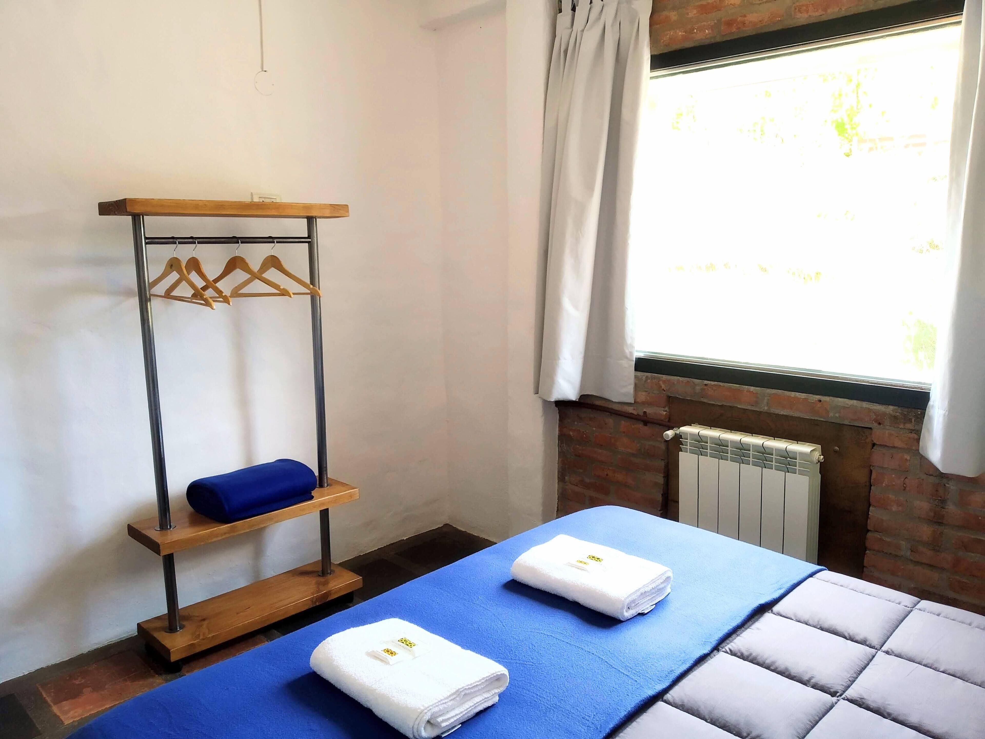 A room with a bed, a blue towel, a white towel, a wooden hanger rack, and a radiator.