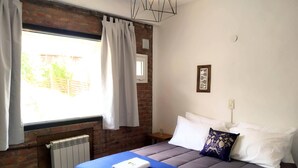 Camera d'albergo con parete in mattoni, finestra con tende, un letto con copripiumino blu, cuscini bianchi e un cuscino blu con motivo floreale.