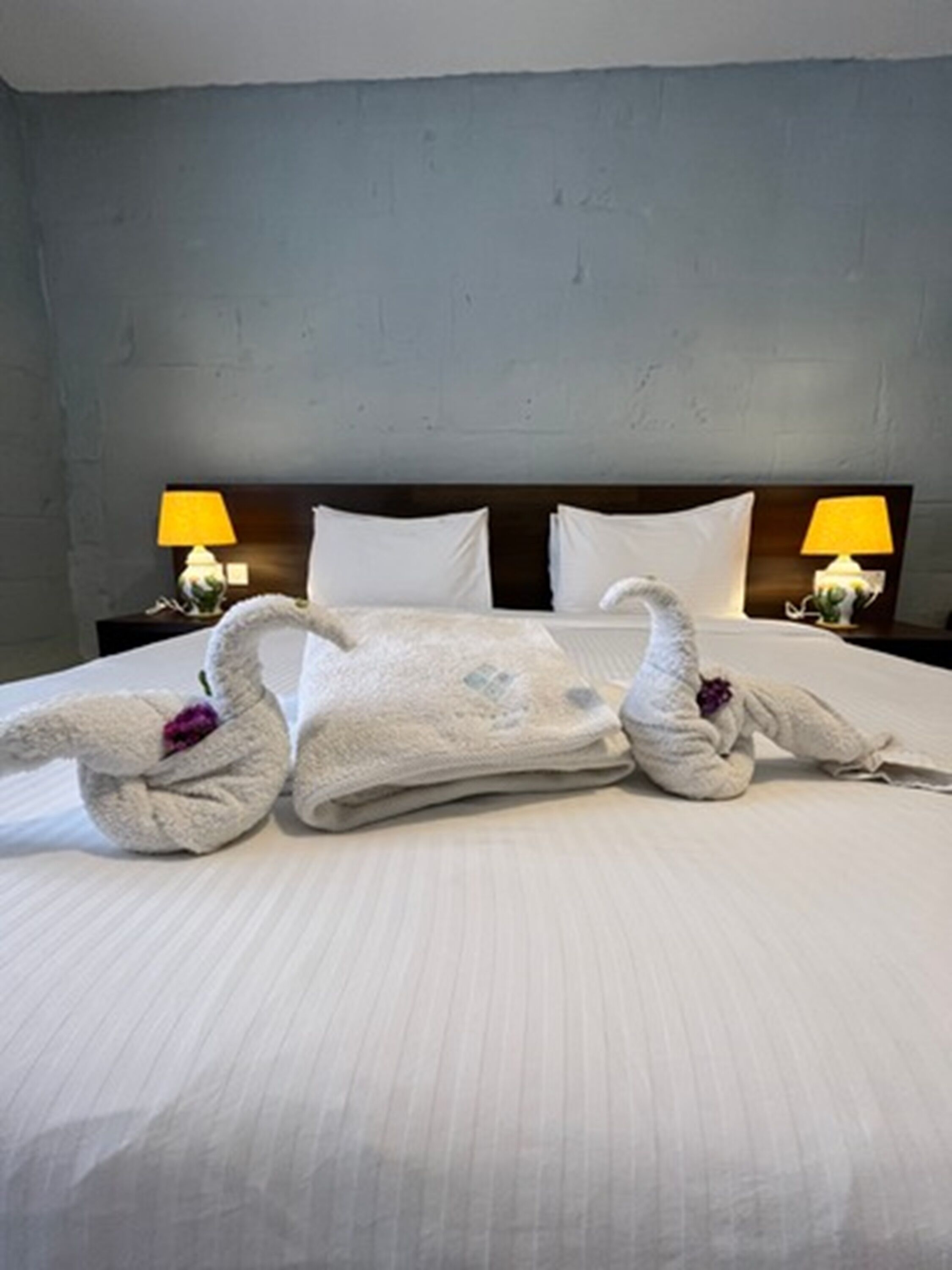 Una cama bien hecha con un diseño de cisne en una toalla, dos lámparas de noche y una pared gris con textura.