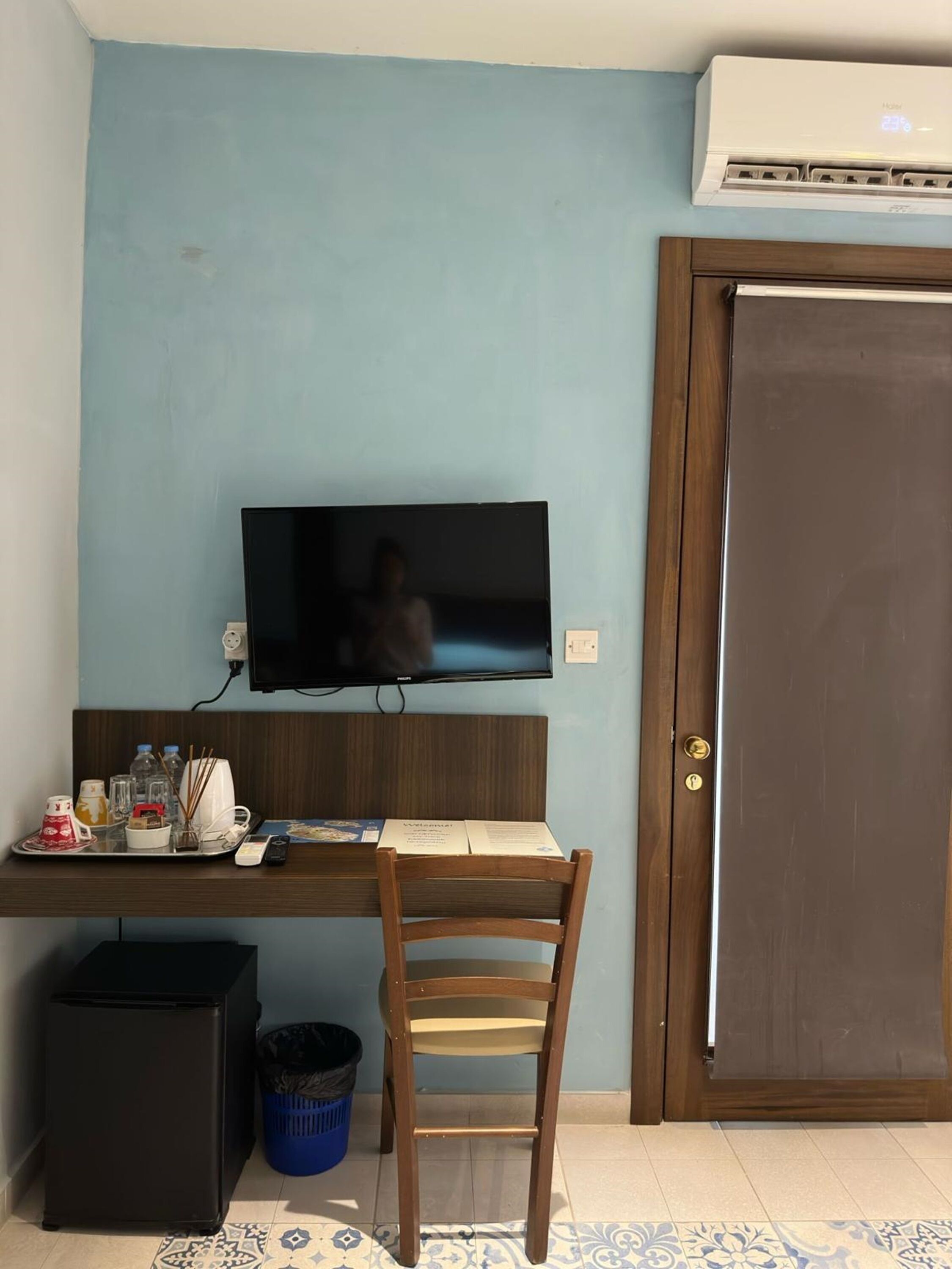Una stanza con una scrivania, una televisione a parete, una porta e un cestino.
