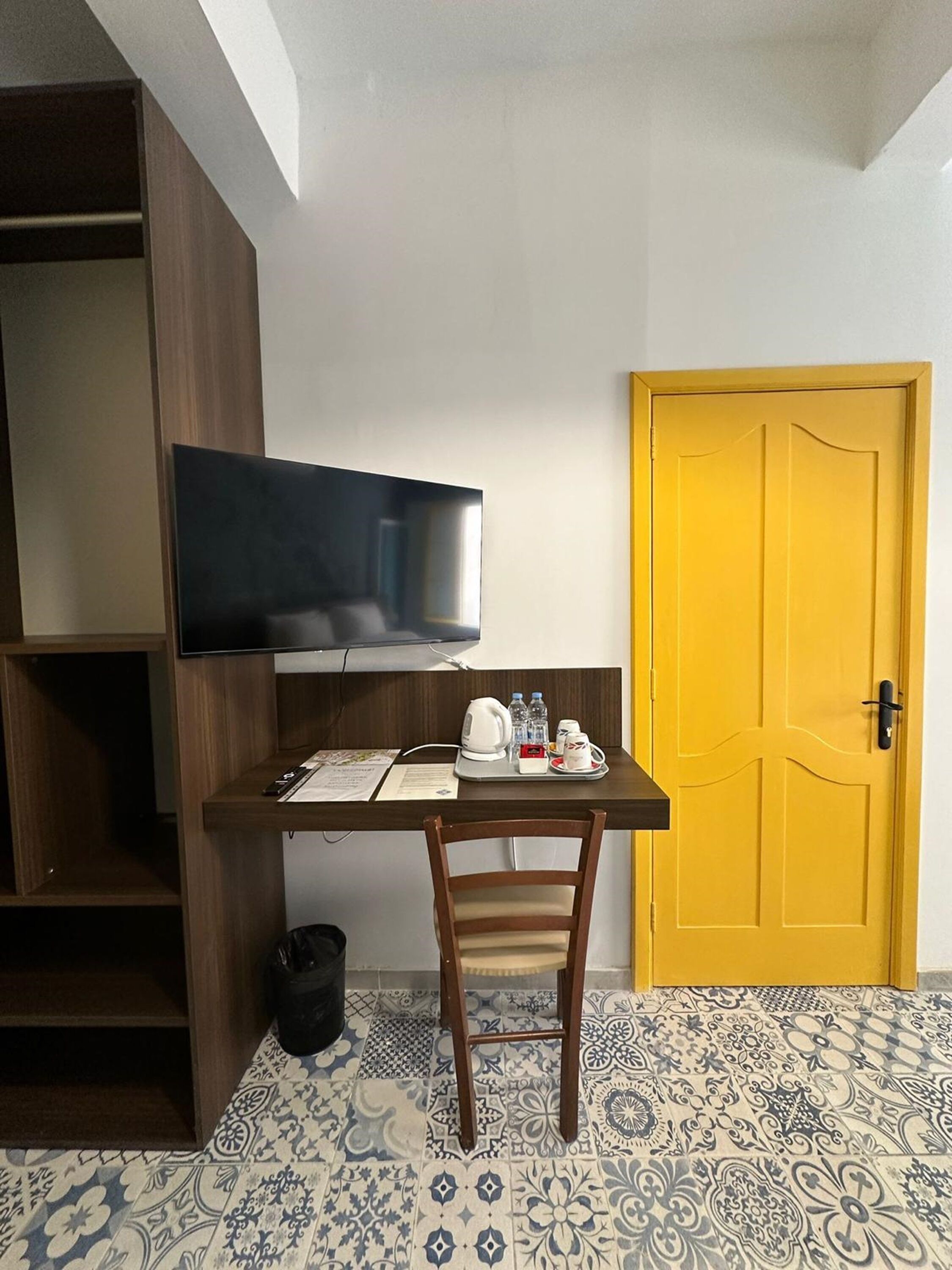 A small room with a wooden desk, a television mounted on the wall, a yellow door, and patterned floor tiles.
