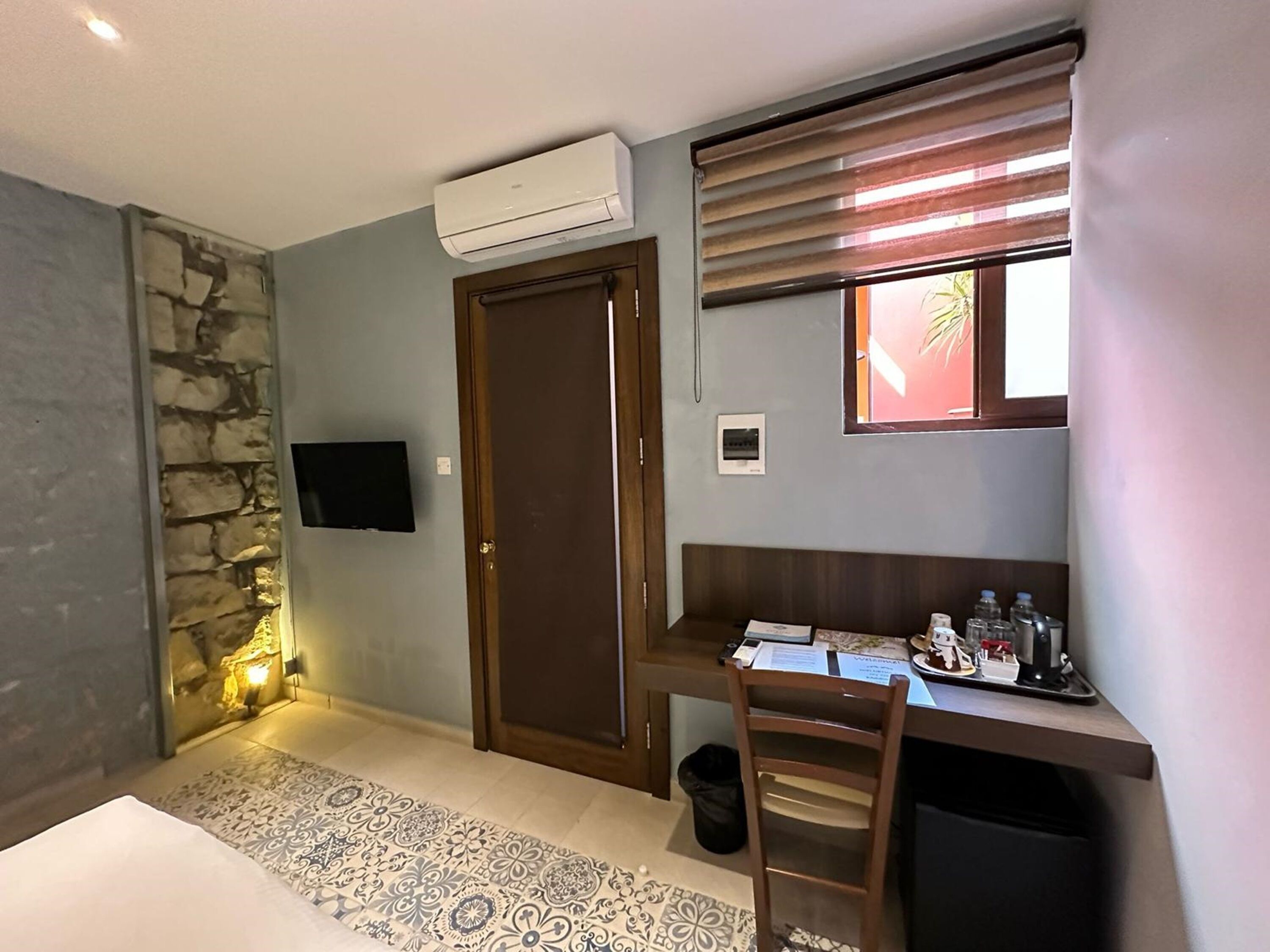 Una habitación con un muro de acento de piedra, un televisor montado en la pared, una puerta de madera, un escritorio con una silla, un cesto de basura pequeño y una ventana con persianas.