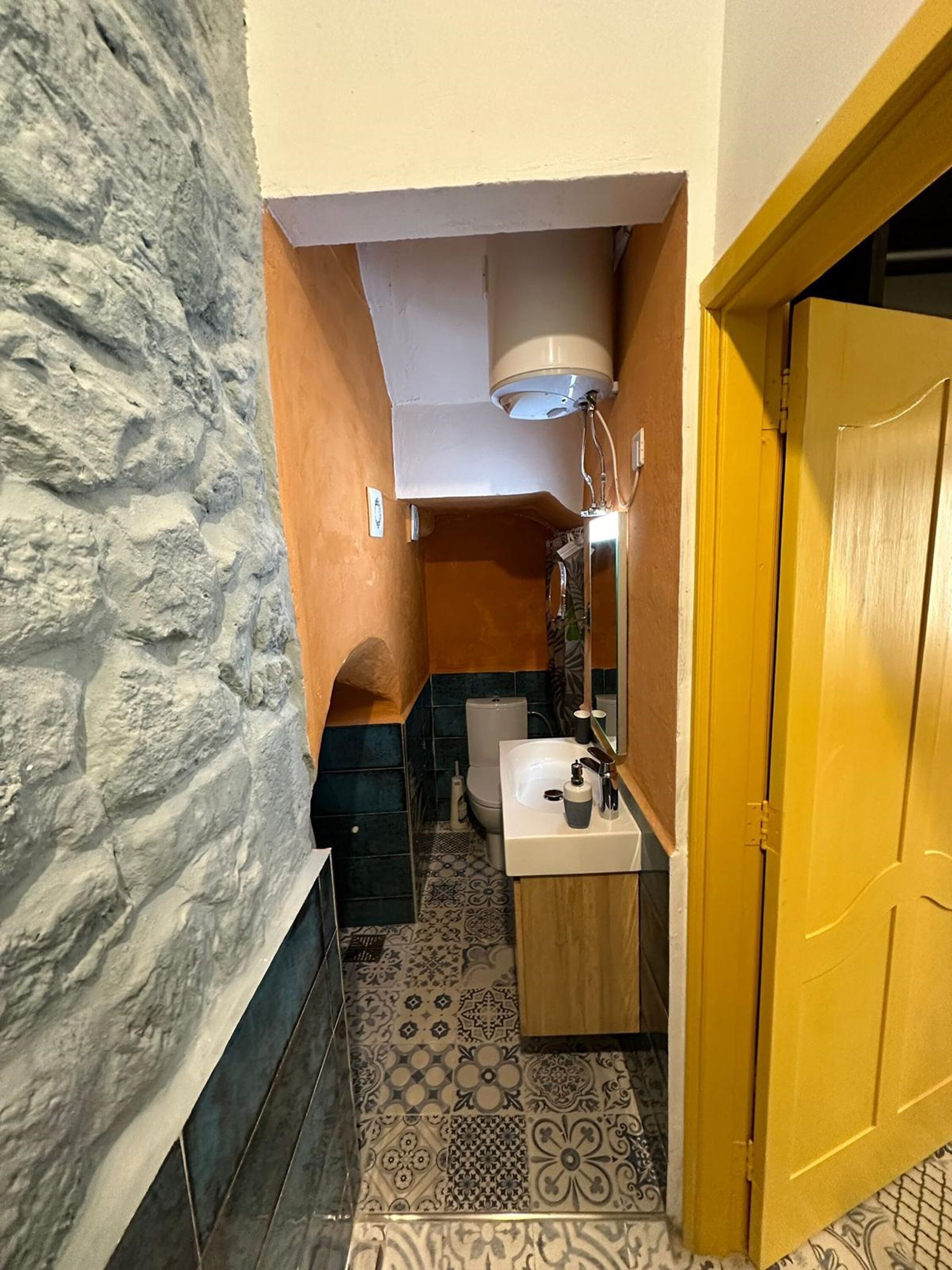 Un baño estrecho con pared de piedra, puerta de madera, lavamanos blanco y baldosas estampadas.