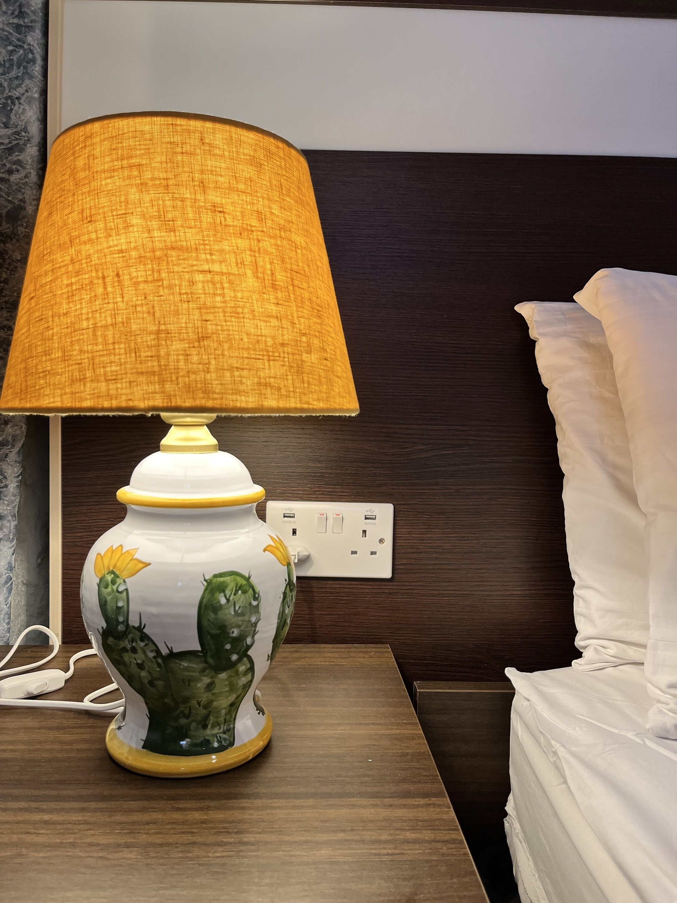Una lámpara con pantalla texturizada sobre una mesita de noche junto a una cama.