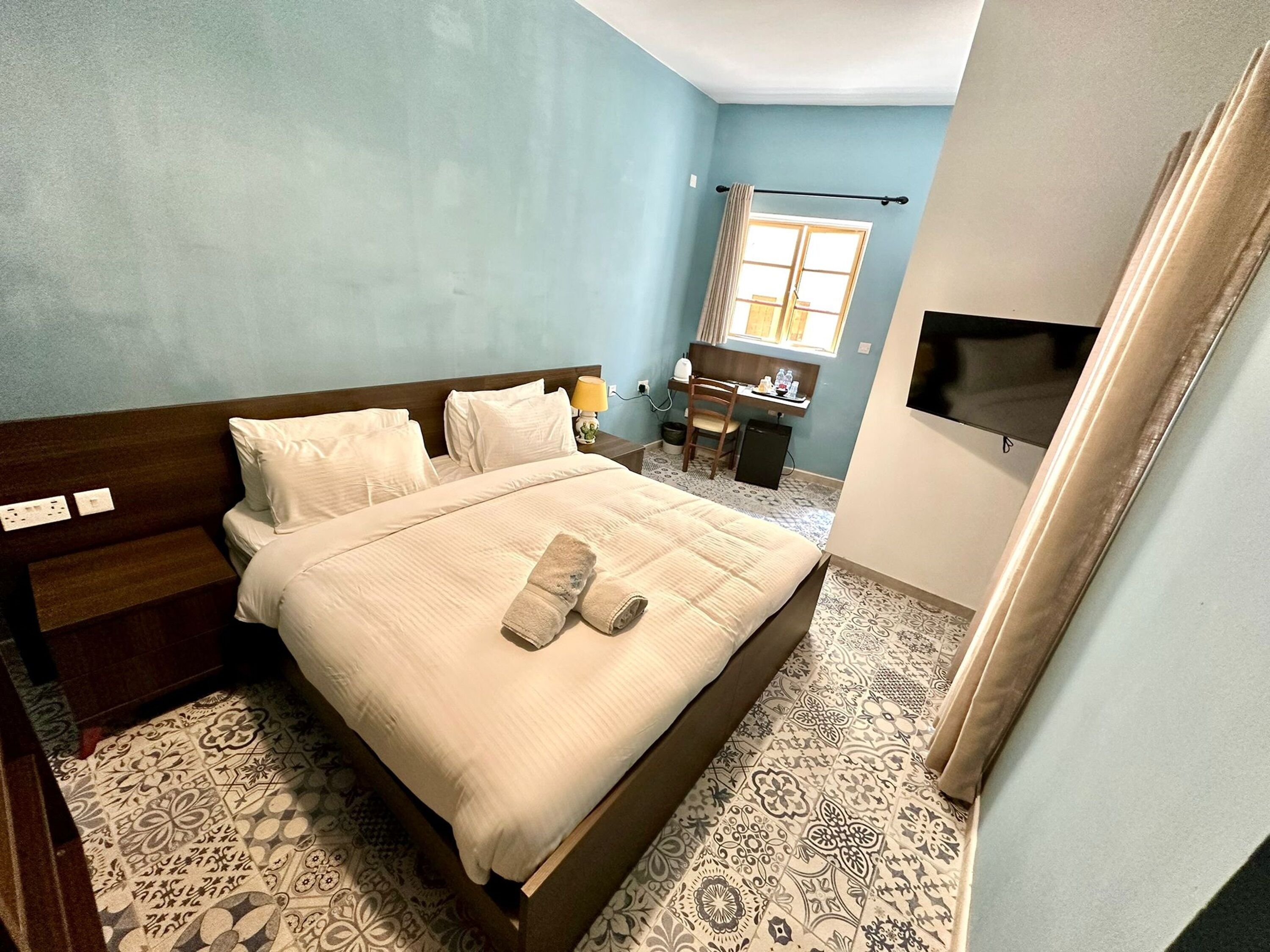 A bedroom with a bed, a desk, a chair, a TV, and patterned flooring.