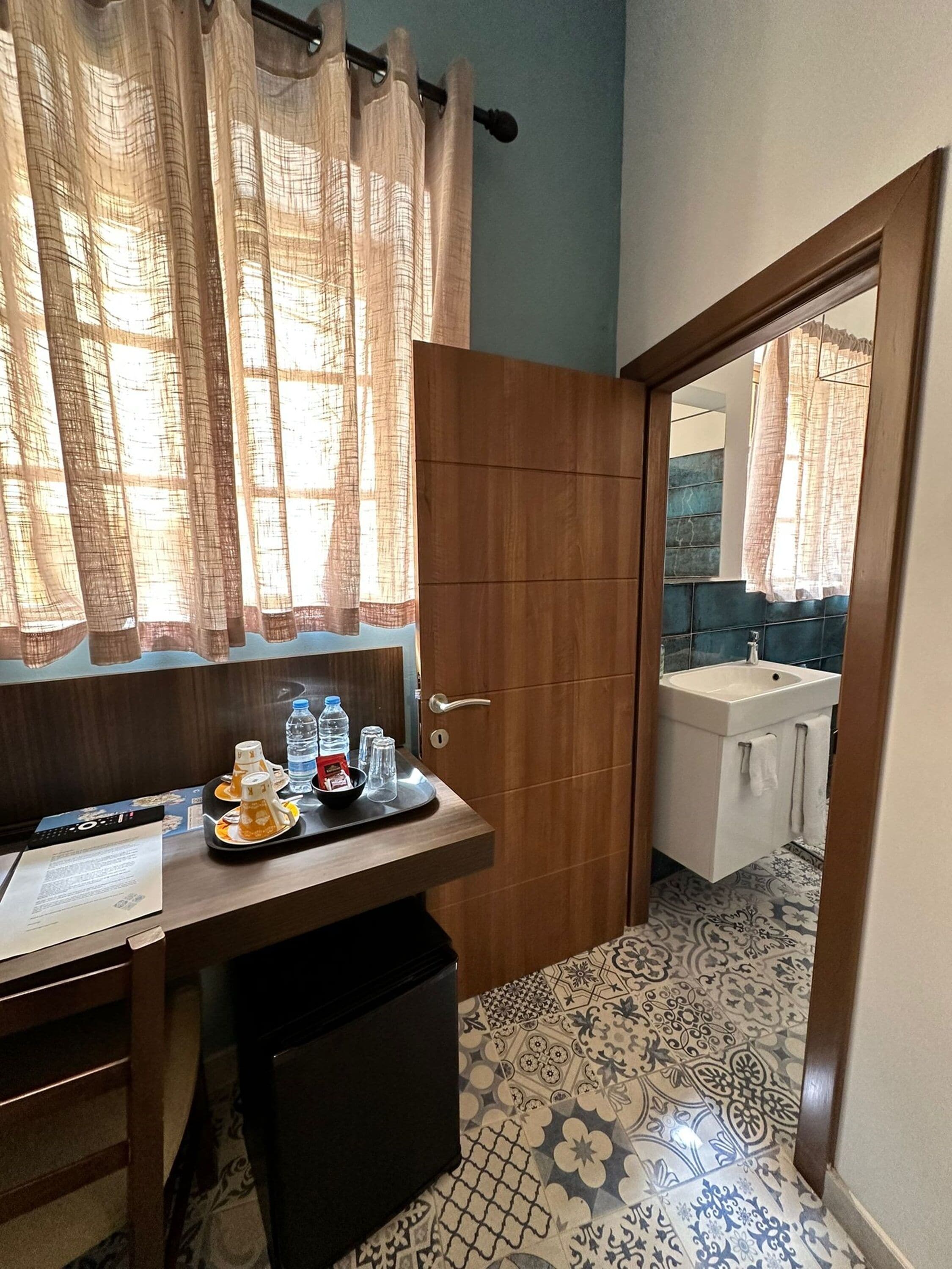 Ein Hotelzimmer mit einem hölzernen Schreibtisch, einer Mülleimer, einer Ablage mit Wasserflaschen und Gläsern, und einer Tür zu einem Badezimmer mit Waschbecken und Spiegel.