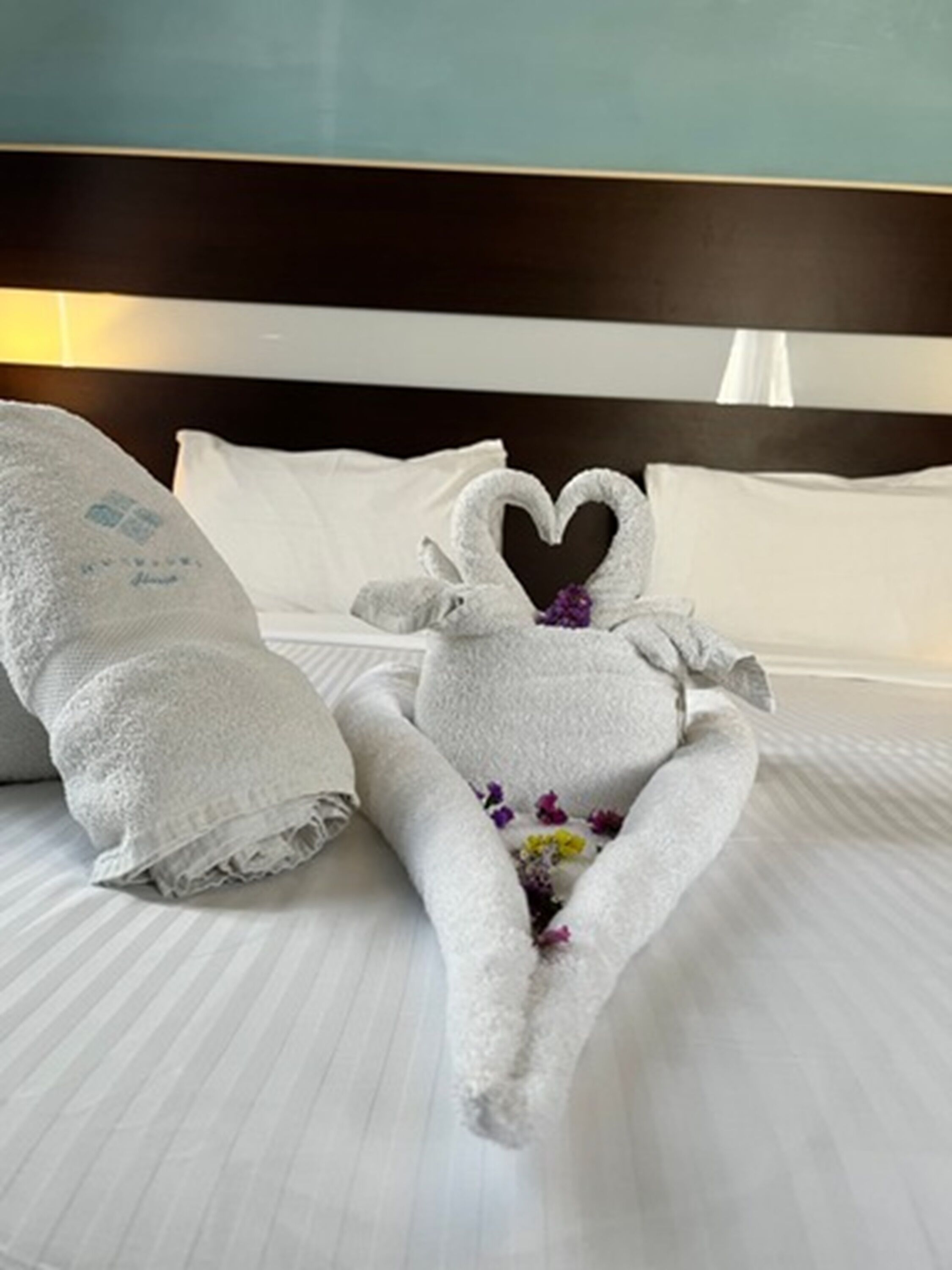 Una cama bien tendida con una toalla dispuesta en forma de corazón y una toalla con diseño floral.