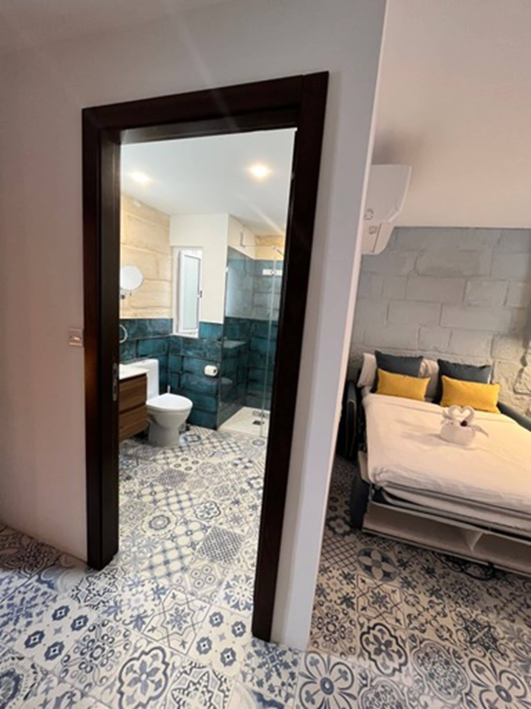 Ein Schlafzimmer mit Bett, Nachttisch und einer Ablage. Durch eine offene Tür ist ein Badezimmer zu sehen, mit Toilette, Waschbecken und Dusche. Der Boden ist mit gemusterten Fliesen belegt.