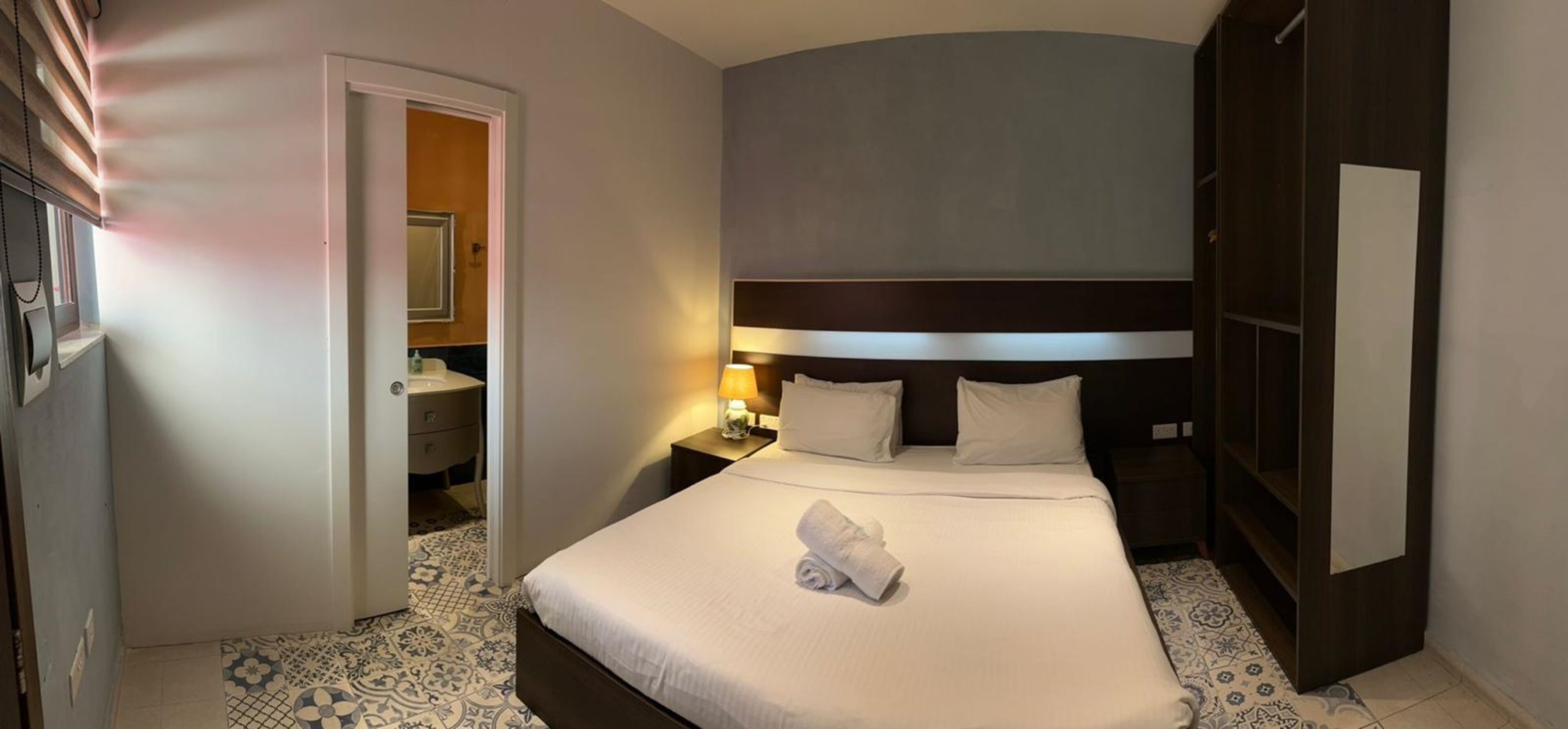 A hotel room with a neatly made bed, a bedside table with a lamp, a wardrobe, and a bathroom visible through an open door.