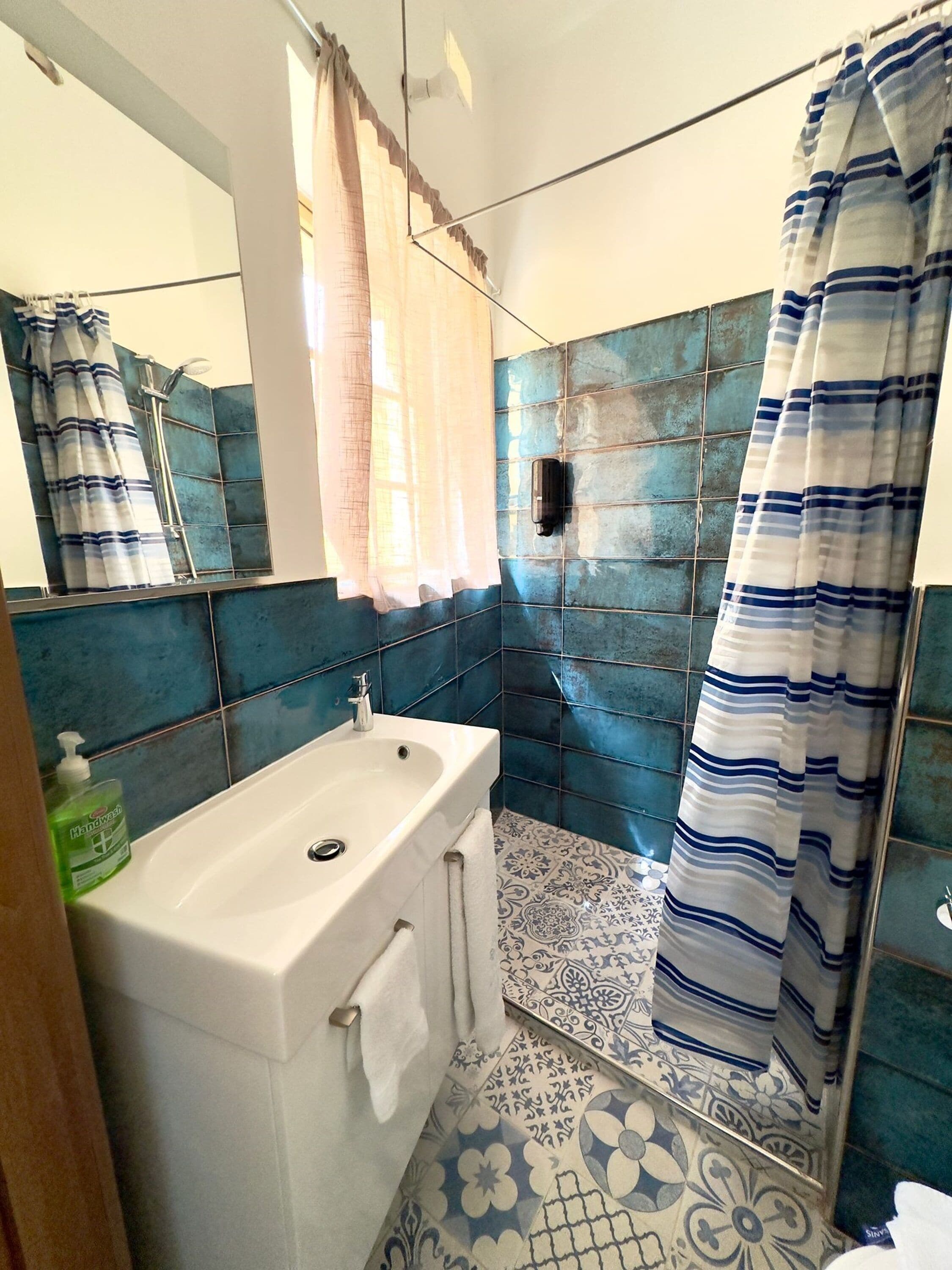Un baño con un lavamanos blanco, azulejos blancos y azules estampados, una cortina de ducha y un espejo.