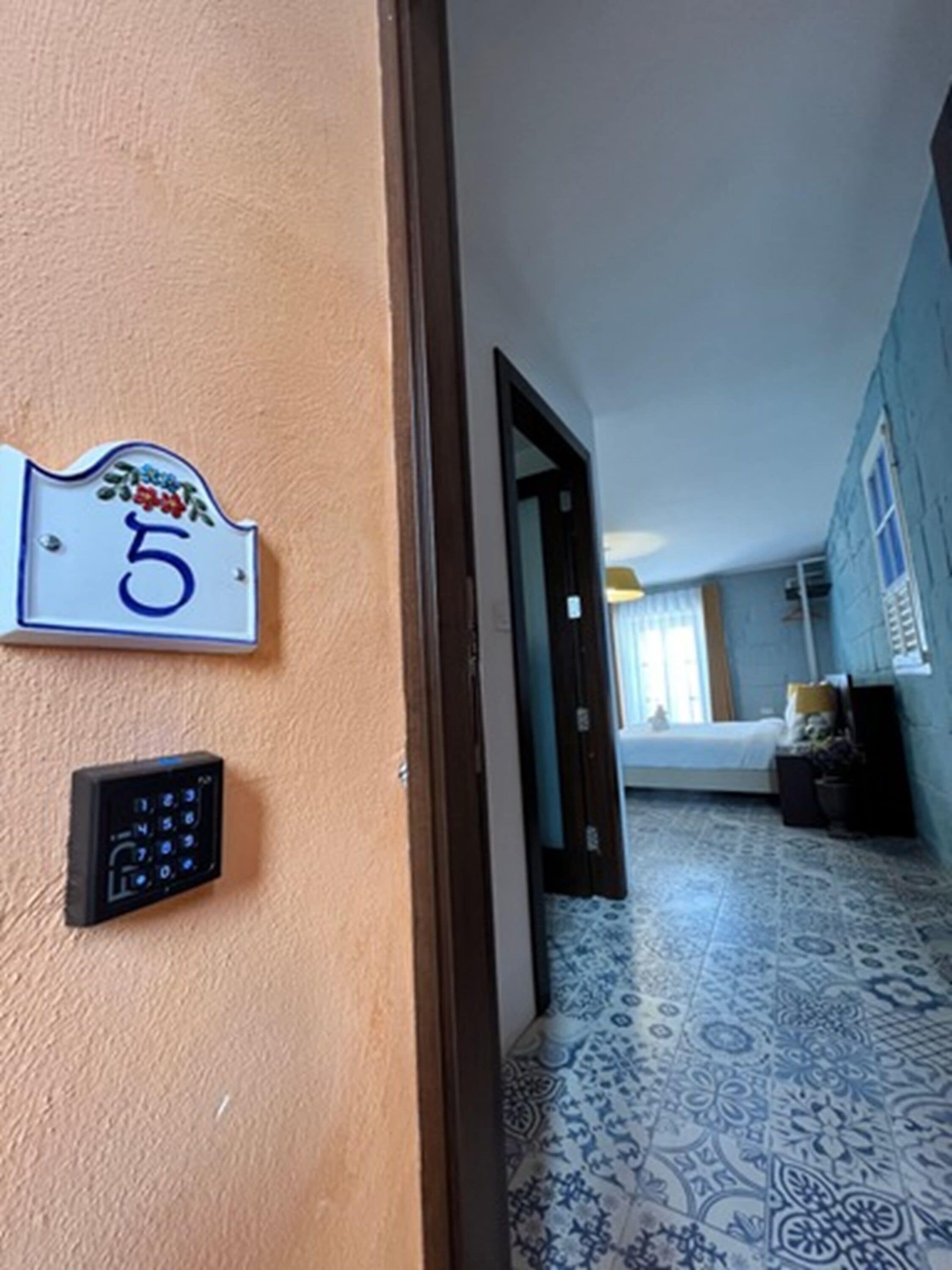 Een gang met een deur die leidt naar een slaapkamer. Een nummerbord is aan de muur bevestigd en een keypad zit op de deur.
