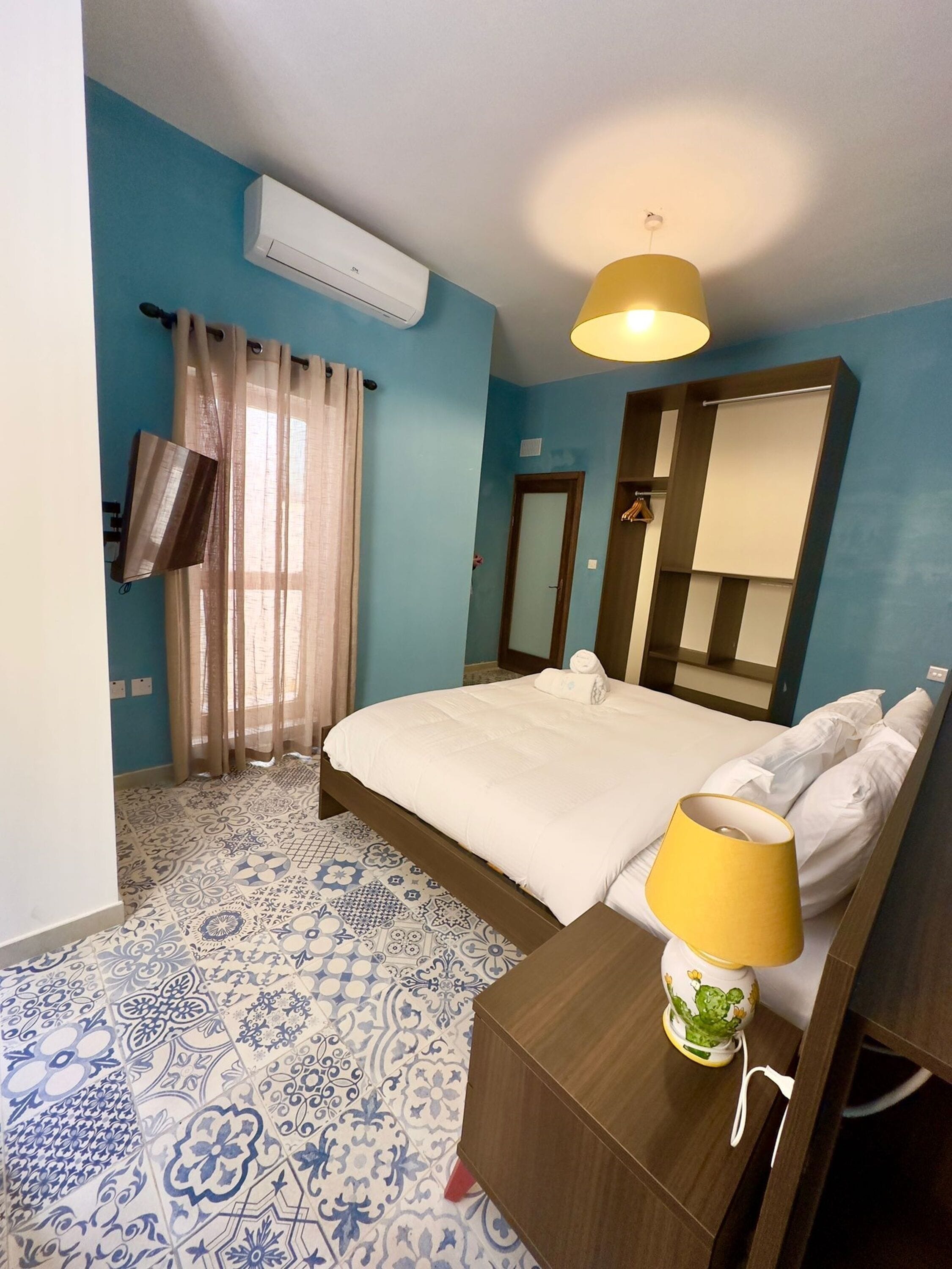 Een hotelkamer met een bed, een bedlampje, een spiegel, een airconditioning en een raam met gordijnen.