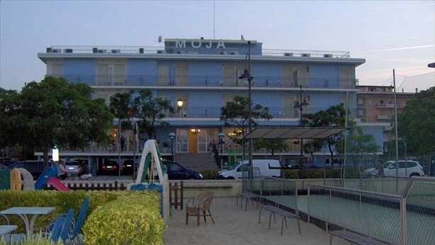 A building named MOJA with a blue facade, outdoor seating area, and a fenced tennis court. - Hotel Moja (Cesenatico)