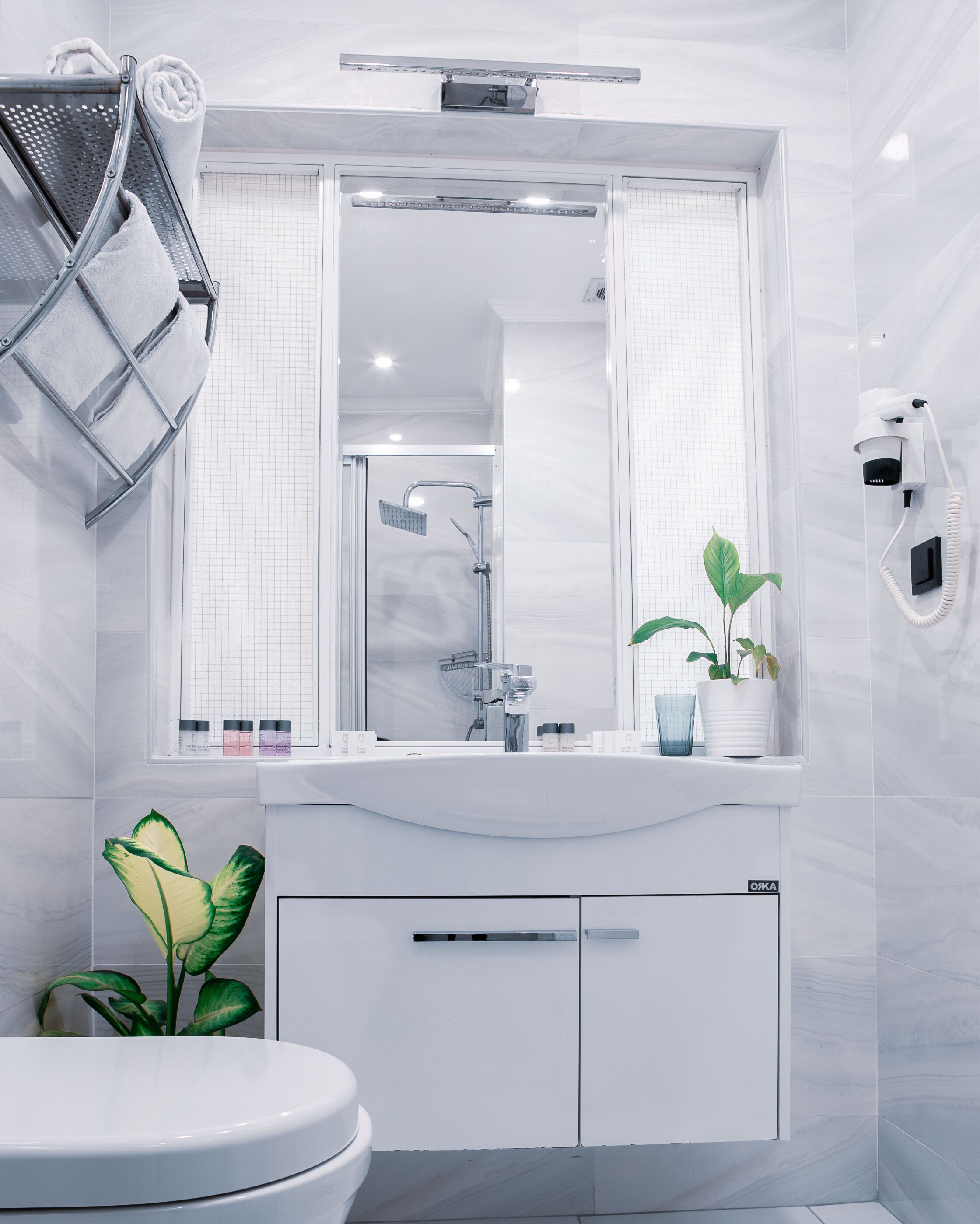 A modern bathroom with a white sink, a mirror, a shower, and a toilet. There are plants on the sink and a towel rack above the mirror.