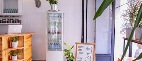 A reception area with a wooden desk, a laptop, a refrigerated display case, and a signboard listing coffee, cake, lunch, and daily fresh items.