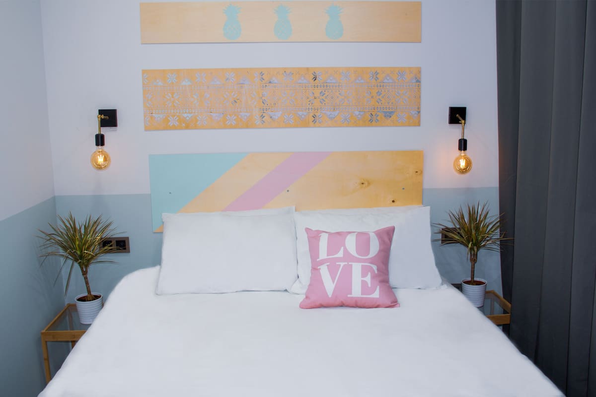 A bedroom with a bed, two potted plants, a wall-mounted light fixture, and a decorative wall piece.